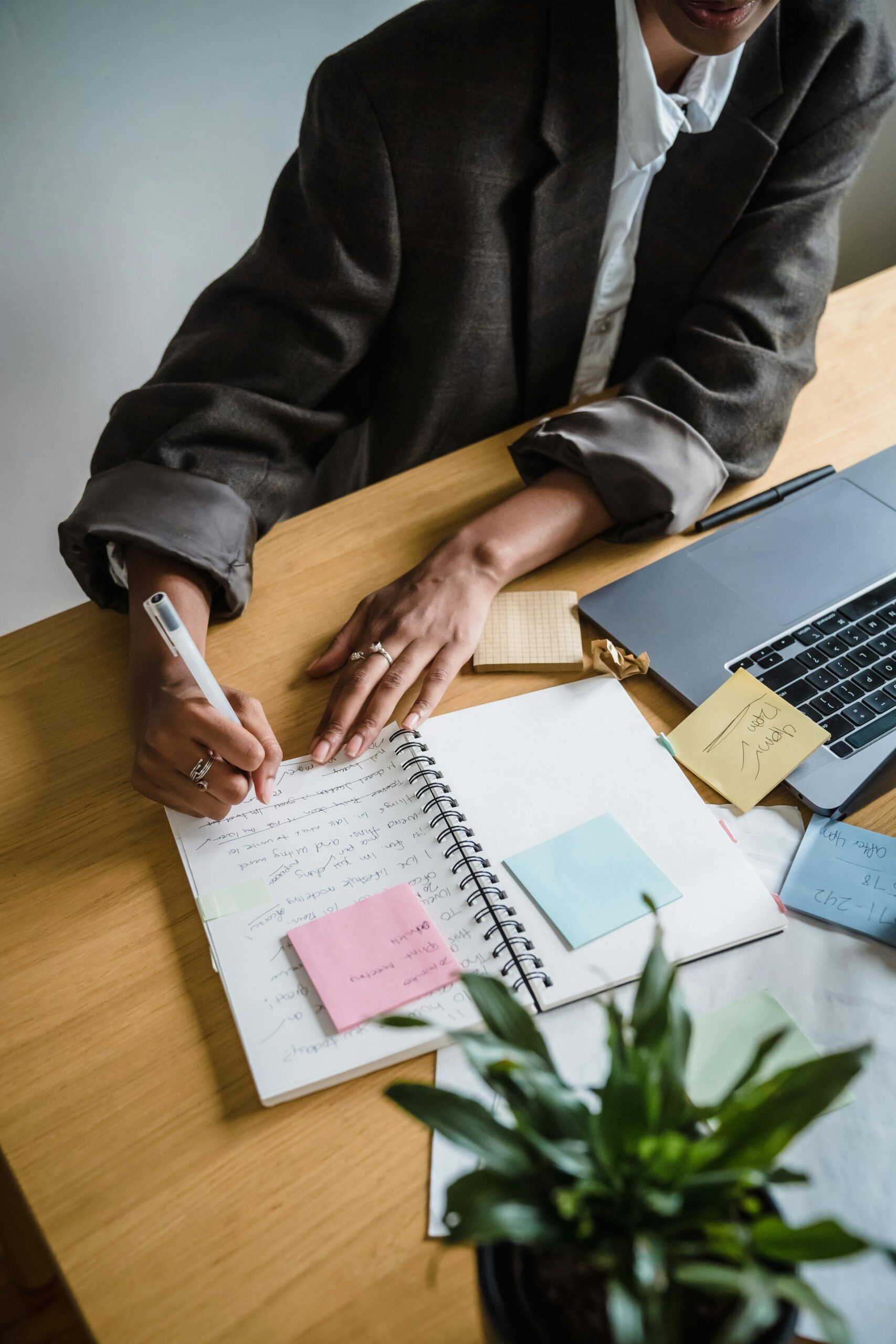 A professional woman writes notes in a notebook at a desk with a laptop and sticky notes.